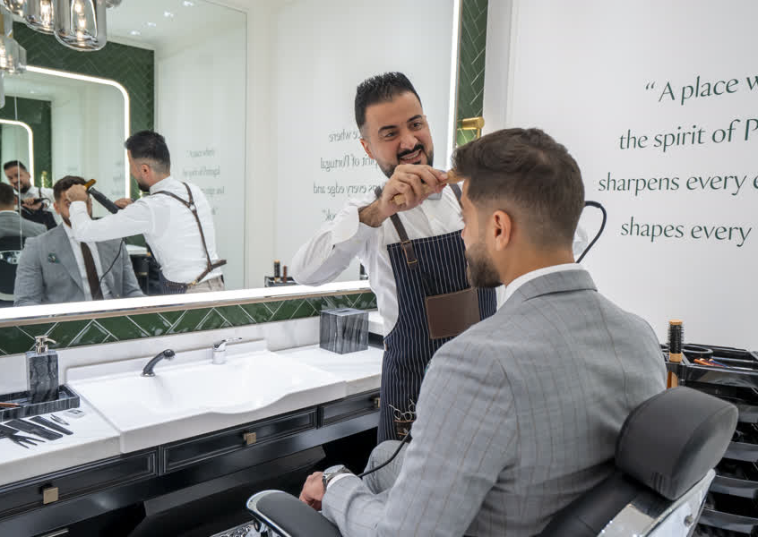 A skilled barber at A Barbearia Portuguesa grooming a client in a tailored grey suit, in a boutique setting with green tiled walls and marble finishes.